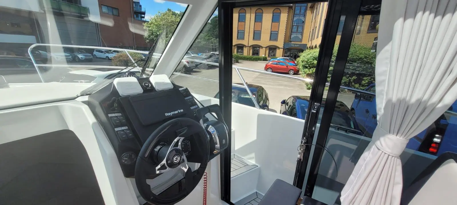 Interior of a boat cabin showing a steering wheel, Raymarine display, and a view of parked cars outside.